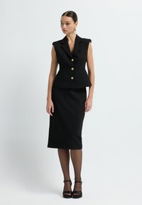 Gilet noir ajusté avec épaules rembourrées et boutons dorés, associé à une jupe crayon noire. Modèle portant des talons aiguilles noirs.
