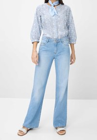 Mujer vistiendo una blusa de estampado azul claro con mangas abullonadas, pantalones de mezclilla azul claro de pierna ancha, sandalias blancas y una bufanda al cuello de color azul claro.