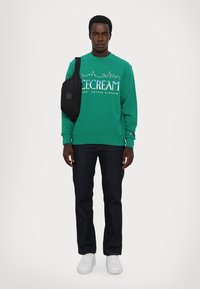 Grøn sweatshirt med "ICECREAM" logo, mørke jeans og hvide sneakers. Modellen bærer en sort crossbody taske. Simpelt design, afslappet pasform.