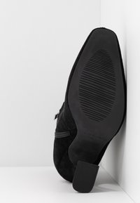 Chaussure à talon noire positionnée verticalement avec une semelle texturée et une fermeture éclair latérale, reposant contre un coin formé par des murs et un sol blancs.