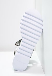 Bílá podešev sandálu s výraznými příčnými drážkami, částečně viditelný pásek s kovovými detaily, na bílém rohovém pozadí.