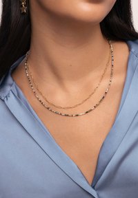 Gros plan sur une personne portant des colliers superposés : une chaîne en or et un collier de perles avec de petites pierres multicolores, avec un col de chemise bleu visible.