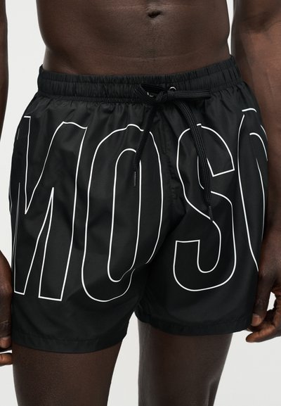 Κοντινό πλάνο ενός ατόμου που φοράει μαύρο μαγιό βερμούδα με μεγάλα λευκά περιγράμματα γραμμάτων και μαύρο κορδόνι στη μέση.