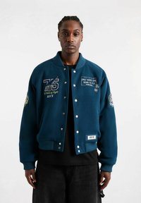 Man in een blauwe varsity jacket met de patches "75 50 Jaar Vliegen" en "Slechts Sommigen Zijn Geboren om te Vliegen", een zwart shirt en zwarte broek.