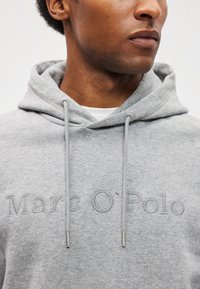 Uomo che indossa un hoodie grigio chiaro di Marc O'Polo con cordini, visibile dalla gola alla parte superiore del petto su uno sfondo chiaro e semplice.