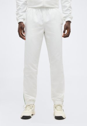 Άτομο που φοράει λευκά παντελόνια jogger με ελαστική μέση και λευκά αθλητικά παπούτσια, στέκεται με τα χέρια χαλαρά στα πλευρά σε απλό φόντο.
