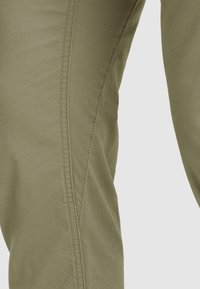 Nahaufnahme von olivgrünen Hosen, die die Stoffstruktur und vertikale Nahtdetails im Oberschenkelbereich vor einem einfarbigen hellen Hintergrund zeigen.