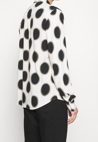 Langarmshirt in Weiß und Schwarz mit großen, kreisförmigen Mustern. Seidige Textur, Kragen und leicht geschwungener Saum. Minimalistisches Design.