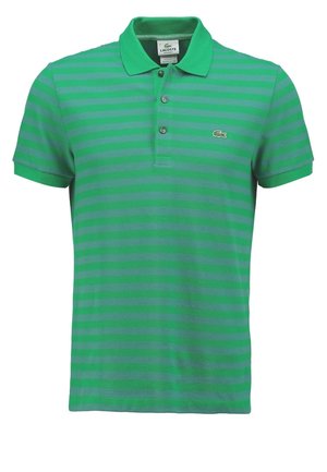 Mand iført grønstribet polo, mørke opkørte jeans, hvide slip-on sko, holder sorte solbriller, står mod en ensfarvet baggrund.