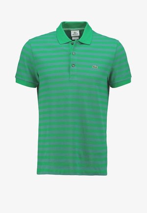 Man met een groen gestreepte polo, donkere opgerolde spijkerbroek, witte instapschoenen, houdt een zwarte zonnebril vast, staand tegen een effen achtergrond.