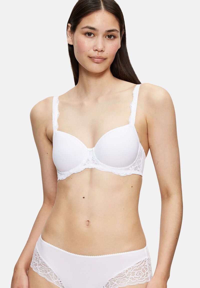 Femme aux longs cheveux foncés portant un soutien-gorge blanc orné de dentelle et une culotte assortie à finition en dentelle, sur un fond uni.