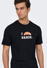 Μαύρο βαμβακερό T-shirt με γραφικό σχέδιο που περιλαμβάνει το κείμενο "I PARIS" και μια εικόνα κρουασάν πάνω από το κείμενο. Μανίκια κοντά.