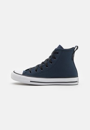 Basket montante en toile bleu foncé avec lacets noirs, bout en caoutchouc blanc et semelle blanche avec des rayures noires sur un fond uni.