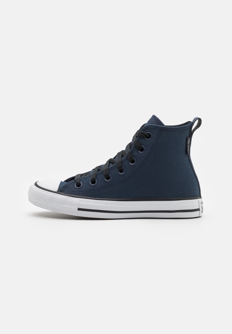 Basket montante en toile bleu foncé avec lacets noirs, bout en caoutchouc blanc et semelle blanche avec des rayures noires sur un fond uni.
