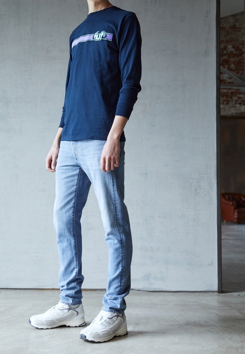 Personne portant une chemise bleu marine à manches longues avec une bande violette, un jean bleu clair et des baskets blanches, debout à l'intérieur sur un sol en béton.
