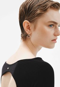 Pendientes en forma de lágrima de plata con un acabado suave, llevados por una persona con el cabello corto, sobre un fondo que resalta una prenda negra.
