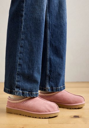 Roze UGG klompen van zacht suède materiaal, met een ronde neus en gestikte rand, gecombineerd met donkerblauwe spijkerbroek, rustend op een houten vloer.
