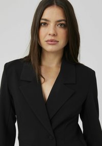 Blazer noir avec un revers cranté, fermeture à un bouton, et une coupe ajustée. Tissu lisse avec une silhouette structurée, peu de détails visibles.