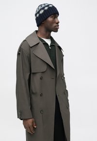 Gray trench coat with double-breasted design, large collar, and shoulder epaulets. Worn over a green zip-up sweater and a navy patterned beanie.