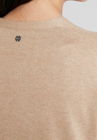 Beige stickad tröja med rund halsringning, ribbade kanter och en liten märkesdetalj vid nackens baksida. Mjuk textur.