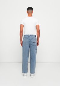 Lichtblauwe denim jeans met een ontspannen pasvorm, voorzien van meerdere zakken en een zwart logo-embleem. Gecombineerd met een nauwsluitend wit T-shirt.