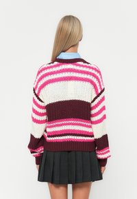 Sweater de malha com riscas brancas, cor-de-rosa e bordô. Corte solto, mangas largas, barra canelada. Combinado com uma saia preta plissada. Vista traseira.