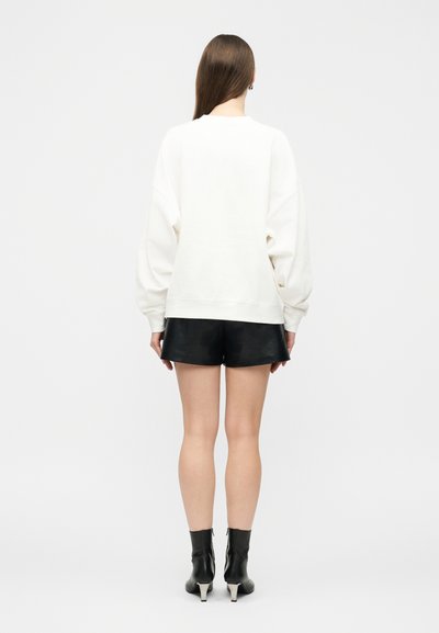 Moteris su ilgais rudos spalvos plaukais, vilkinti baltą per didelį džemperį, juodus odinius šortus ir juodus aukštakulnius aulinukus, stovinti atsukta nugara.