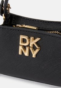 Czarna torebka o zauważalnej fakturze, z metalowym logo "DKNY" w kolorze złotym, zamknięciem na zamek błyskawiczny oraz cienkim paskiem do noszenia.