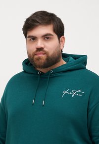 Felpa color turchese realizzata in tessuto morbido, con cappuccio regolabile tramite coulisse, caratterizzata da un logo bianco in corsivo sul petto e una vestibilità comoda.