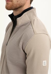 Beżowa kurtka zapinana na zamek, z teksturowanym materiałem na ramionach i klatce piersiowej, a gładkim wykończeniem na rękawach. Naszywka z logo na ramieniu.