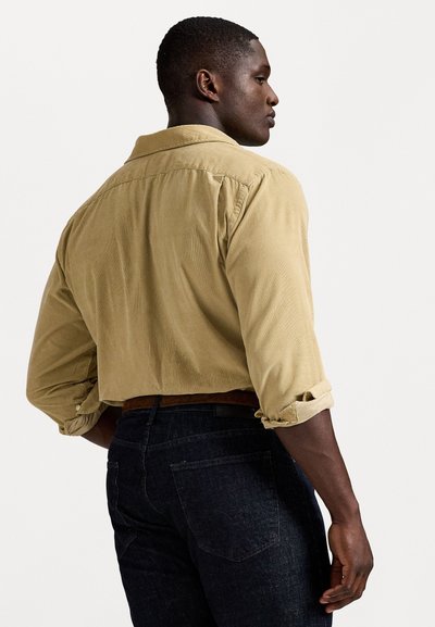 Homem de perfil, vestindo uma camisa bege de manga longa com as mangas enroladas e jeans azul-escuro com um cinto castanho, contra um fundo branco.