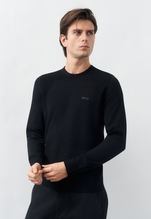 Jeune homme aux cheveux bruns portant un t-shirt à manches longues noir ajusté avec le logo "BOSS", debout devant un fond gris clair uni.