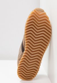 Semelle de chaussure en caoutchouc épais de couleur beige avec un motif de bande de roulement en zigzag large, inclinée contre un mur blanc et une surface.