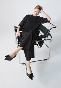T-shirt oversize nera e pantaloni a gamba larga abbinati a scarpe slingback nere lucide con punte affusolate, poggiati su una sedia in cromo.