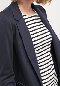 Marinblå kavaj i strukturerad tyg som matchas med en vit och svart randig skjorta. Kavajen har slagkrage och skräddarsydd passform.