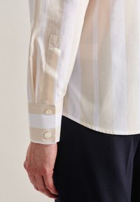 Man's left arm wearing a beige and white striped long-sleeve shirt with buttoned cuff, resting beside dark trousers.