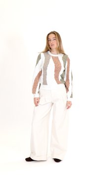Sieviete stāv, valkājot baltu džemperi ar bēšām, brūnām un olīvu abstraktām svītrām, platām baltām biksēm un tumši brūnām kurpēm.