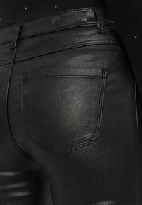 Črne, sijoče skinny hlače z gladko teksturo, ki imajo edinstveno obliko žepov in oznako na hrbtni strani pasu.