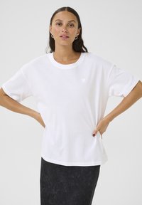 T-shirt en coton blanc à manches courtes, coupe décontractée, avec un petit motif en forme de cœur sur la poitrine gauche. Associé à une jupe texturée foncée.