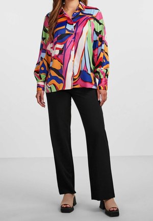 Femme portant une chemise à manches longues à boutons avec un imprimé abstrait coloré, un pantalon large noir et des talons compensés noirs à bout ouvert.