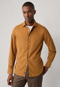 Camisa de pana color mostaza de manga larga con botón en la parte delantera, textura suave y una sutil etiqueta de la marca en el dobladillo.