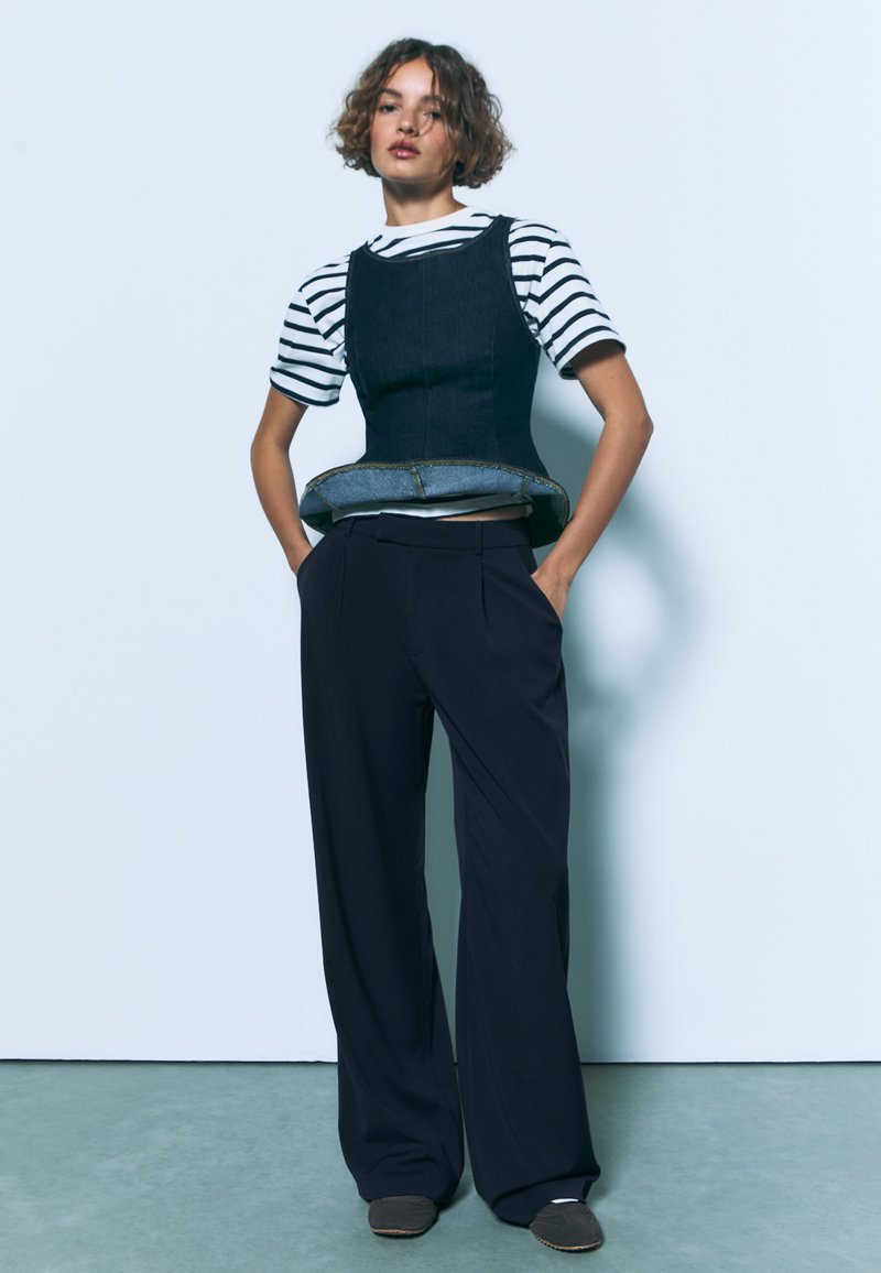 Denim top in korsetstijl over een zwart-wit gestreept shirt, gecombineerd met zwarte broek met wijde pijpen en grijze instapschoenen. Aardetinten achtergrond.