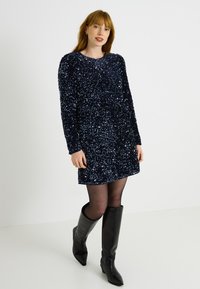 Vestido azul marino de lentejuelas con mangas largas, cuello redondo y cintura ceñida, combinado con botas negras hasta la rodilla y medias negras transparentes.