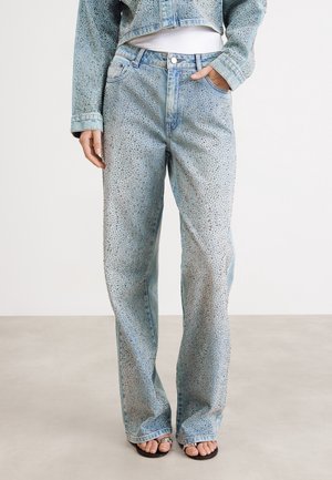 Jasnoniebieskie dżinsy z denimu ozdobione srebrnymi kryształkami na całej powierzchni, w zestawie z dopasowaną kurtką dżinsową również wysadzaną kryształkami oraz czarnymi sandałami z odkrytymi palcami.