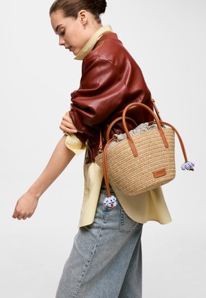 Joven mujer con chaqueta de cuero marrón y vaqueros claros lleva un bolso de mimbre tejido con asas de cuero y una bolsa de tela con cordón.