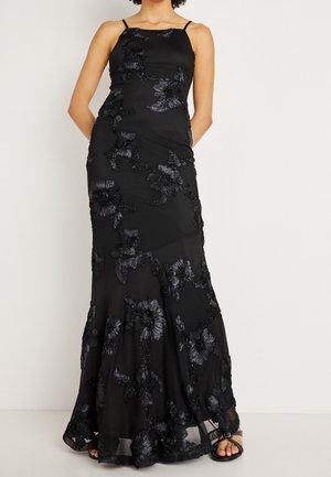 Mujer con un vestido negro sin mangas con bordados florales y tirantes finos, posando frente a un fondo liso y claro.