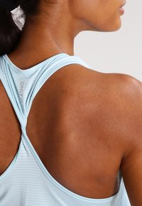 Jasnoniebieski sportowy top z krzyżującym się tyłem, ozdobiony delikatnymi poziomymi paskami i gładką fakturą, podkreślający krzywizny ramion i pleców.