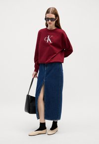 Felpa bordeaux con logo, abbinata a una gonna di jeans con spacco frontale, calze nere e scarpe beige senza lacci. Inclusa una borsa a tracolla nera.