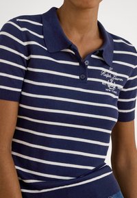 Persona, kas valkā tumši zilu ar baltām svītrām īsroku polo kreklu ar izšūtu tekstu un emblēmu uz kreisās krūšu puses.