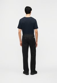 Άνδρας που στέκεται με την πλάτη προς το φακό, φορώντας κοντομάνικο μπλε ναυτικό πουκάμισο, μαύρο παντελόνι cargo και μαύρα αθλητικά παπούτσια σε απλό φόντο.